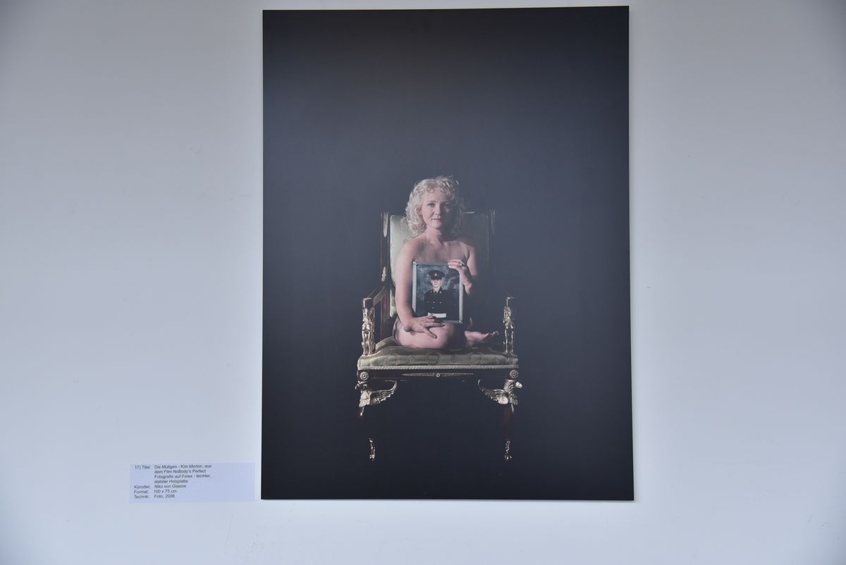 Ein Bild der Galerie, es zeigt eine nackte Frau mit einer Behinderung sitzend auf einem barocken Stuhl, die ein Foto eines Soldaten in Uniform vor sich hält